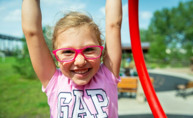 A young child wearing glasses and smiling as she raises her arms up overhead