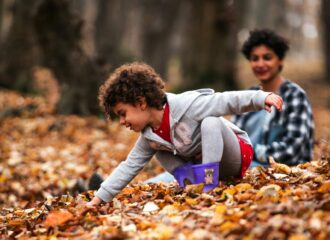 Young child smiling and reaching into dry autumn leaves as an adult looks on in the background